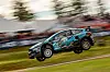 En rallycrossbil i lufta over en grusbane med fartsspor i bakgrunnen.